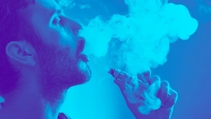 A man exhales a cloud of vapour while holding an electronic device, with a blue-tinted background