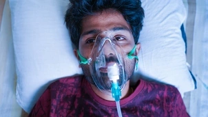 A man lying on a hospital bed wears an oxygen mask, looking upwards